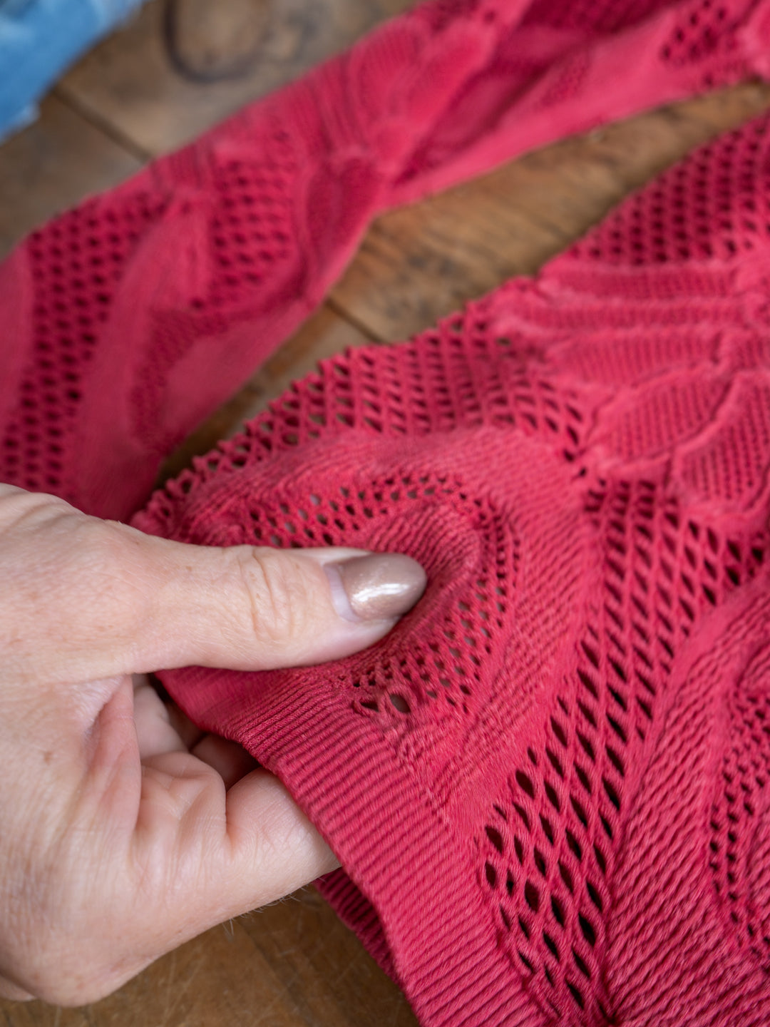 A hand with light nail polish touches The Lexa, a raspberry red crochet top with a small hole in the sheer fabric by Savannah Sevens western life{&}style, displayed on a wooden surface.