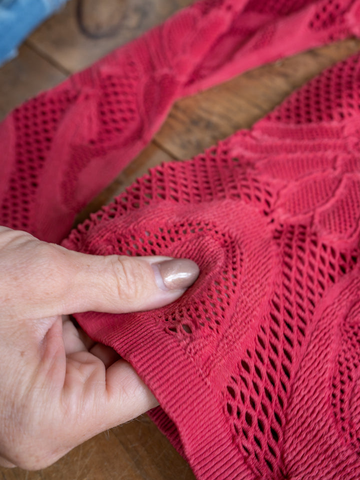 A hand with light nail polish touches The Lexa, a raspberry red crochet top with a small hole in the sheer fabric by Savannah Sevens western life{&}style, displayed on a wooden surface.