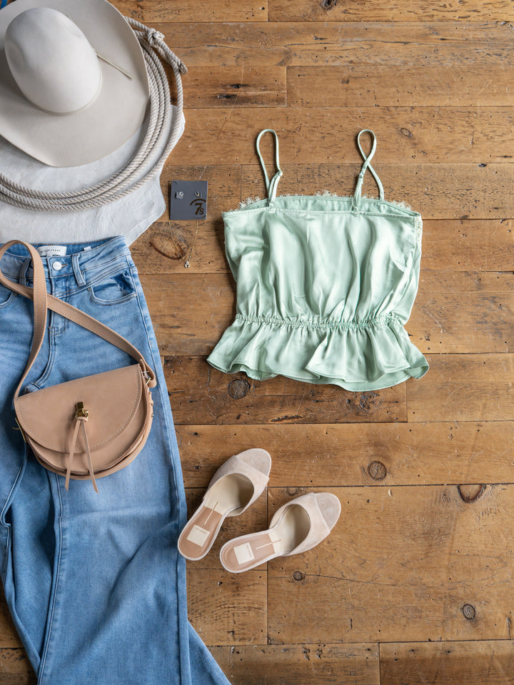 Flat lay featuring The Swansea by Savannah Sevens western life{&}style: white wide-brim hat, light green satin sleeveless top with adjustable straps, light blue jeans, beige crossbody bag, and beige slip-on sandals on a wooden floor.