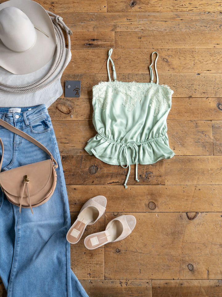 Flat lay of a casual outfit featuring The Swansea light green satin sleeveless top with lace overlay and adjustable straps by Savannah Sevens western life{&}style, styled with light wash jeans, beige flats, a beige crossbody bag, wide-brimmed hat, and round sunglasses.