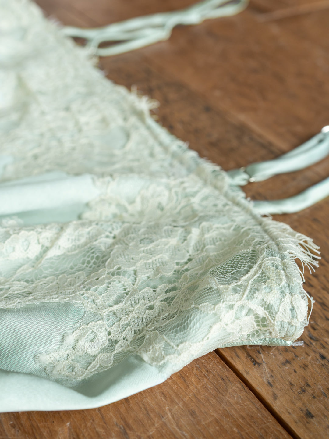 A close-up of The Swansea bralette by Savannah Sevens western life{&}style in light green lace with adjustable straps, displayed on a wooden surface. The delicate lace overlay and soft fabric stand out.