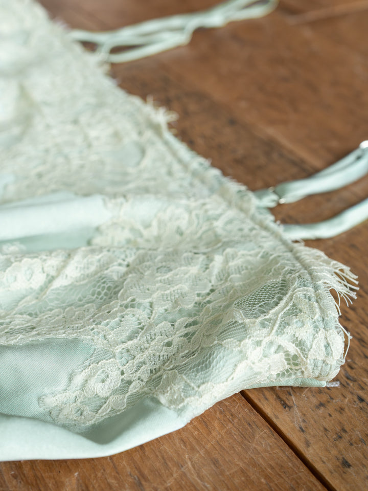 A close-up of The Swansea bralette by Savannah Sevens western life{&}style in light green lace with adjustable straps, displayed on a wooden surface. The delicate lace overlay and soft fabric stand out.