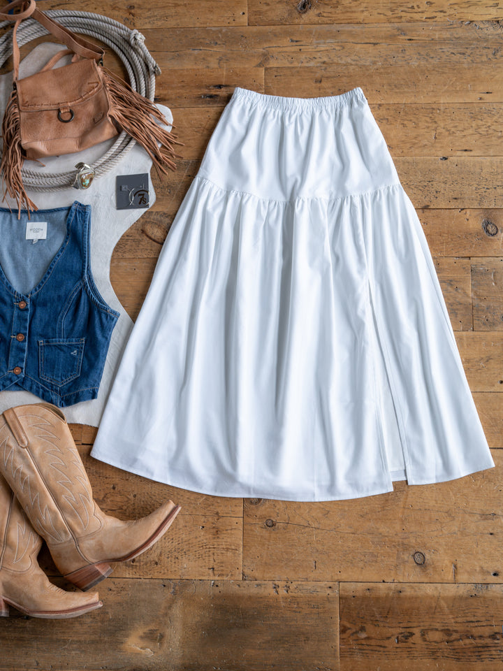 The Estero by Savannah Sevens western life{&}style, a white high-waisted midi skirt with an elastic waistband, is laid flat on wood beside tan cowboy boots, a denim vest, a brown fringed bag, and a turquoise necklace.