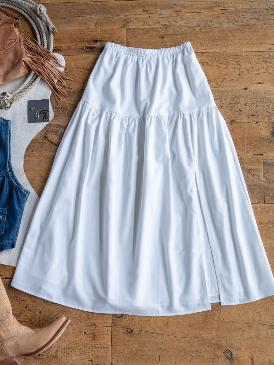 The Estero by Savannah Sevens western life{&}style is a long white high-waisted midi skirt with an elastic waistband, displayed on wood among brown cowboy boots, a fringed bag, denim pieces, and a coiled rope.