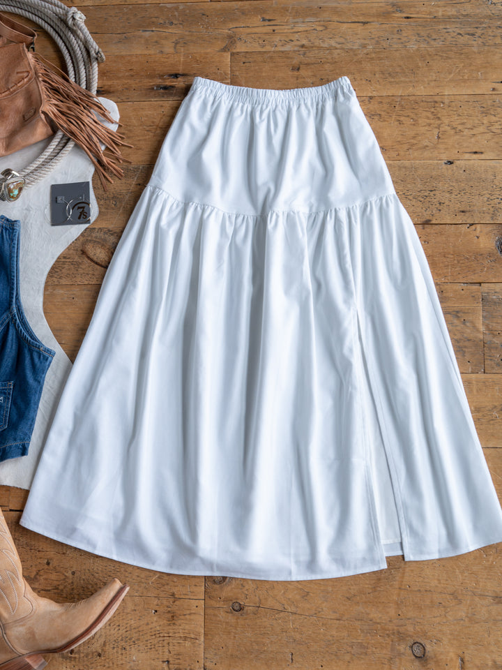 The Estero by Savannah Sevens western life{&}style is a long white high-waisted midi skirt with an elastic waistband, displayed on wood among brown cowboy boots, a fringed bag, denim pieces, and a coiled rope.