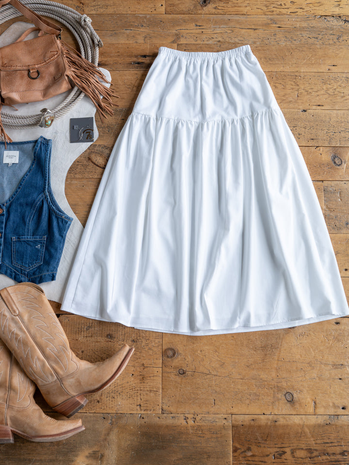 The Estero by Savannah Sevens western life{&}style, a white high waisted midi skirt, is styled on a wooden floor with tan cowboy boots, a brown fringed bag, gray top, blue denim vest, and jewelry.