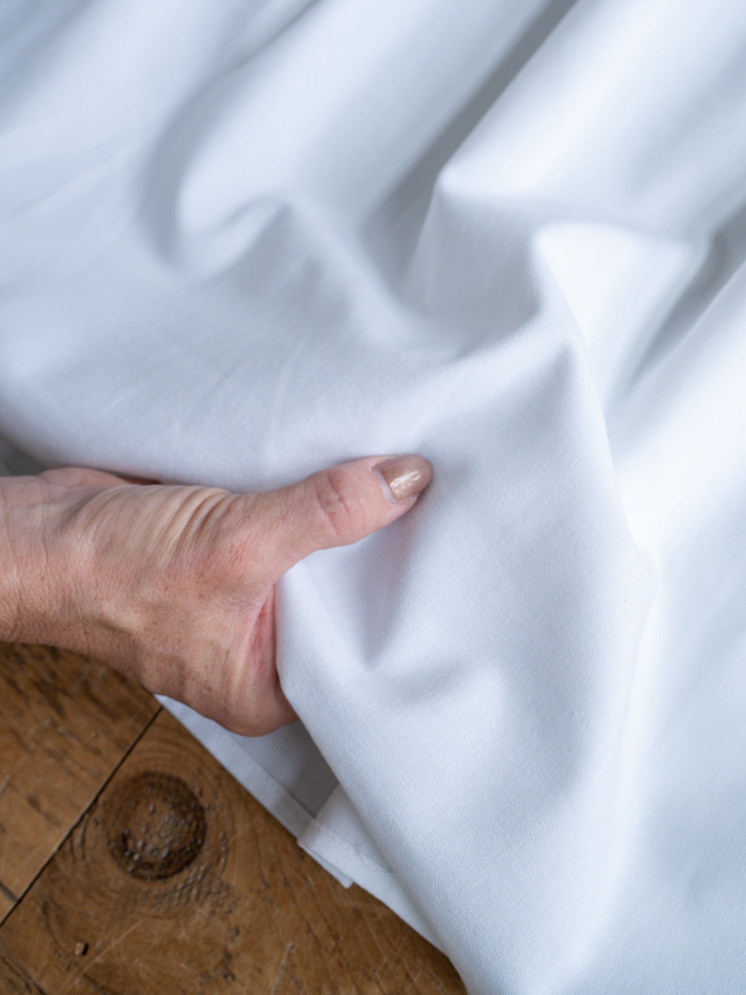A hand with manicured nails gently folds The Estero, a white high waisted midi skirt from Savannah Sevens western life{&}style, on a wooden surface.