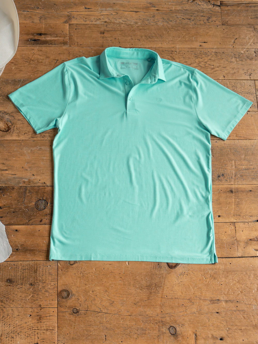 The Griffin Ariat Men's Turquoise Polo by Savannah Sevens western life{&}style features a collared, button-up design, moisture-wicking fabric, and added sun protection—perfect for all-day comfort. It’s shown laid flat on a rustic wooden floor.