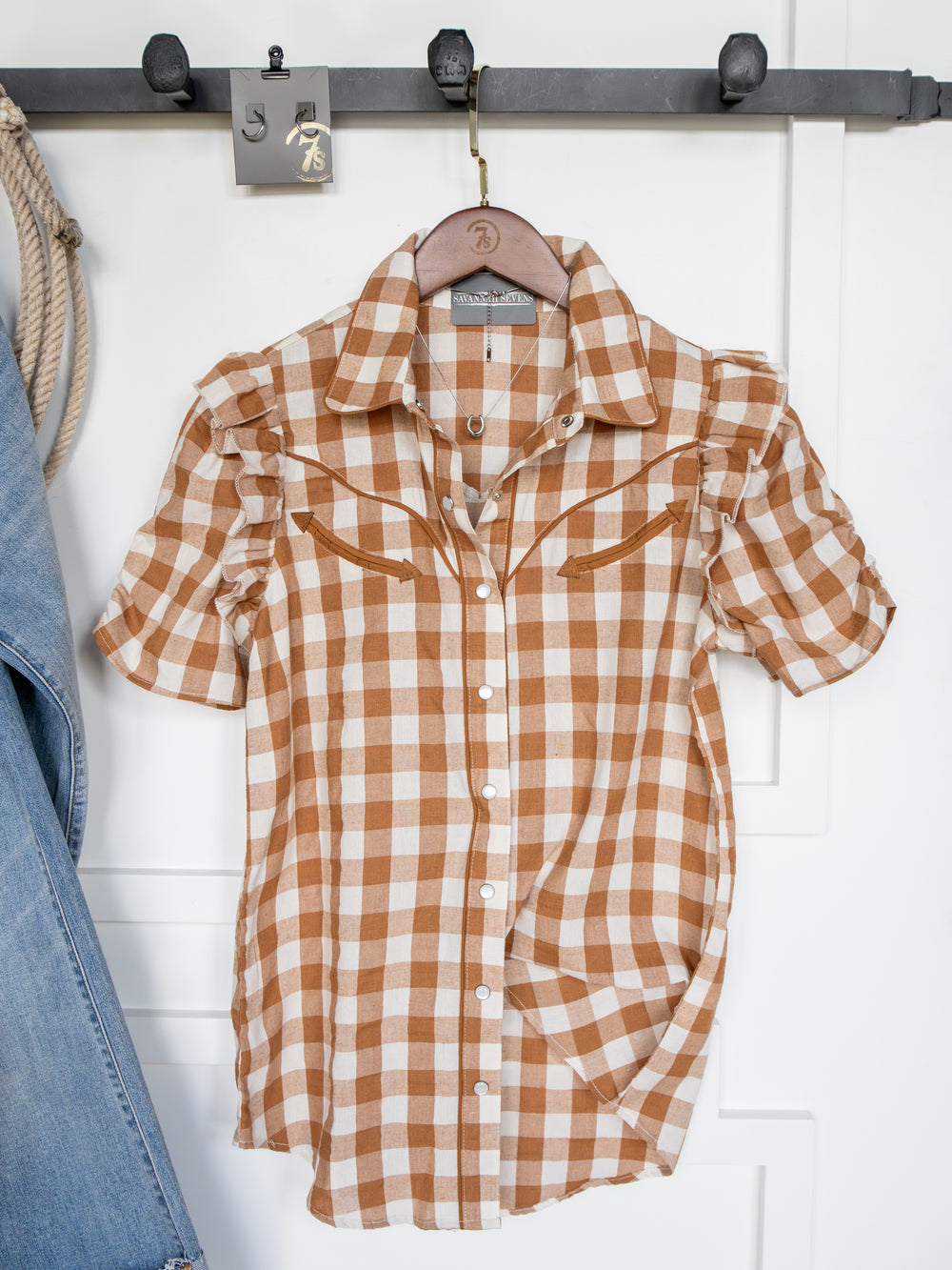The Somona Brown Gingham Pearl Snap Top by Savannah Sevens western life{&}style, featuring short sleeves, hangs on a wooden hanger with blue jeans partially visible to the left.