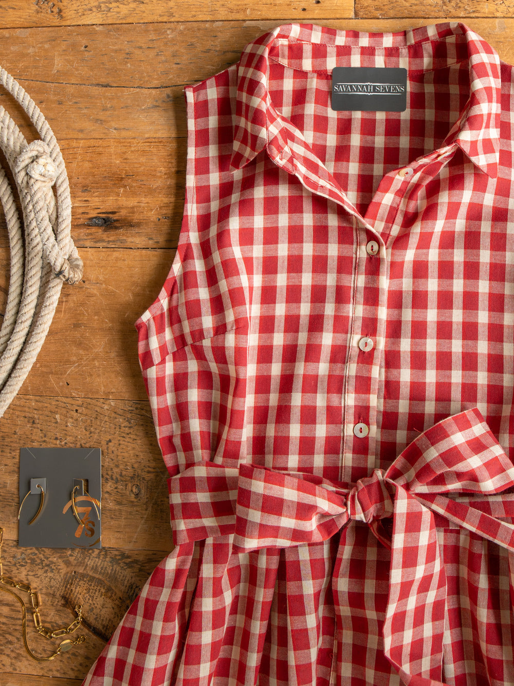 The Tolland Red Gingham Button Up Dress by Savannah Sevens western life{&}style, a sleeveless red-and-white dress with a tied waist, is displayed on wood beside gold earrings, a bracelet, and a coiled rope.