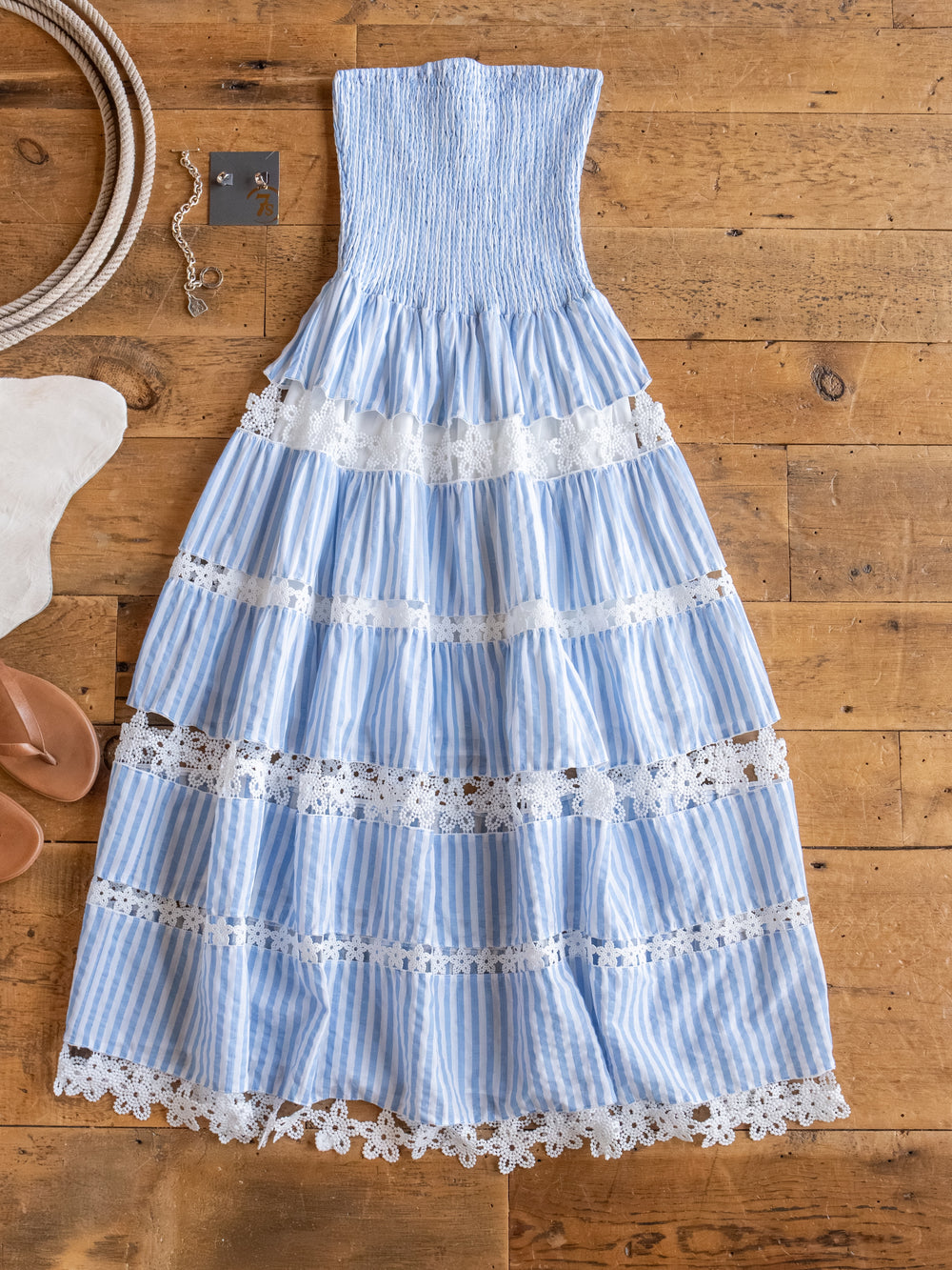 The Lantana Blue Striped Strapless Dress by Savannah Sevens western life{&}style is laid flat on a wooden floor, surrounded by a bracelet, sandals, and a hat.