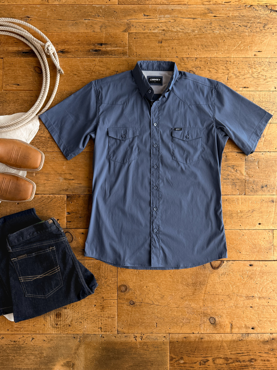 The Weatherford men's short sleeve button up by Savannah Sevens western life&style is displayed on a wooden floor with rope, tan boots, and dark jeans. Its athletic fit combines comfort and style for any occasion.