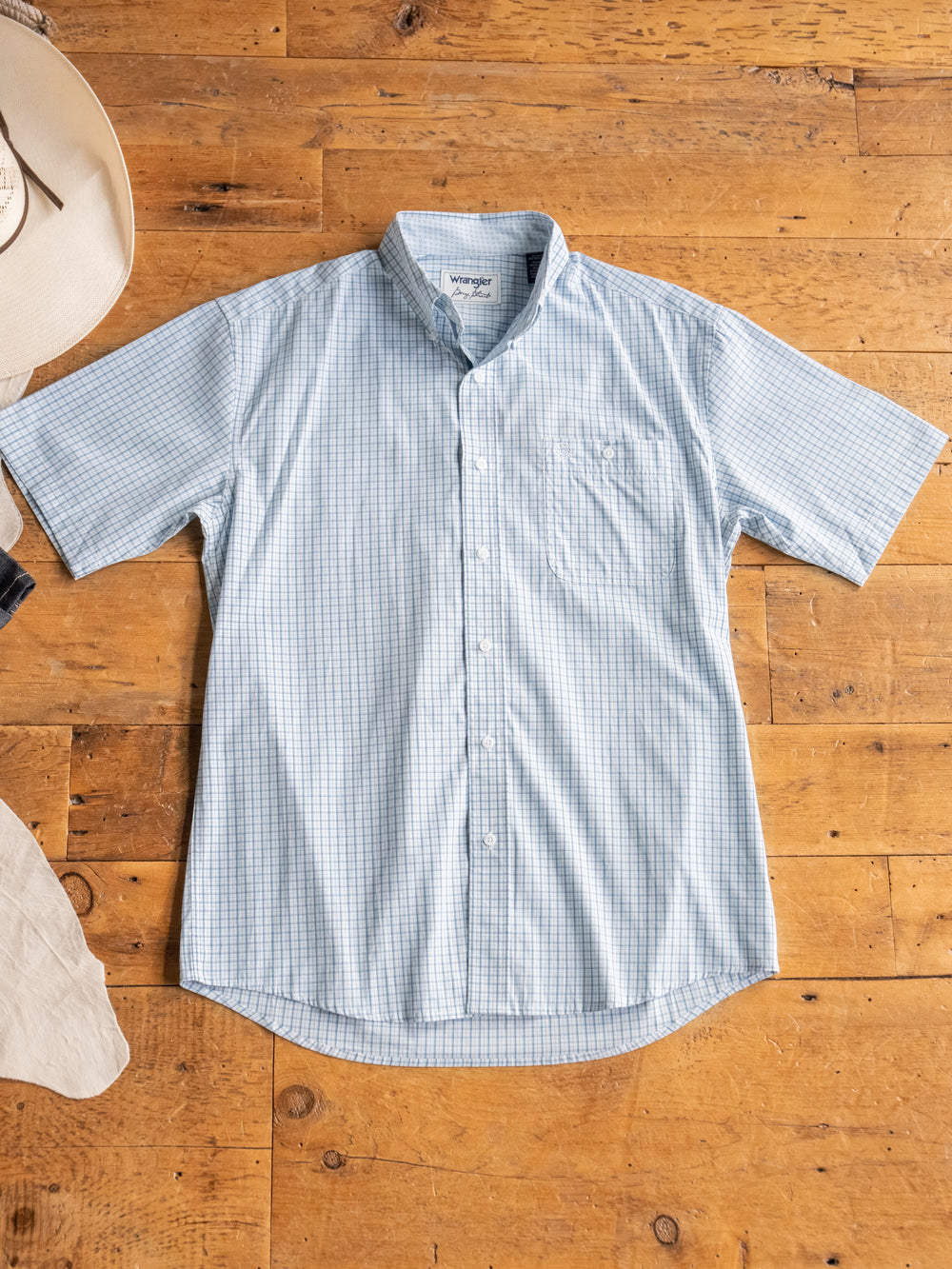 A light blue men's short sleeve button up, The Bentonville from Savannah Sevens western life&style, features a subtle check pattern and is laid flat on a wooden floor with a beige cowboy hat and light-colored fabric beside it.