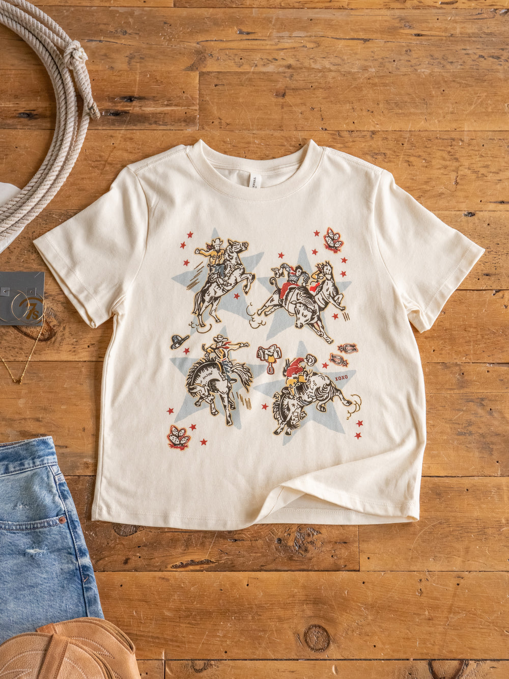 The Star Cowboy tee by Savannah Sevens western life{&}style, a cream 100% cotton shirt with cowboy, red star, and floral graphics, is displayed flat on wood alongside denim shorts and a tan hat.