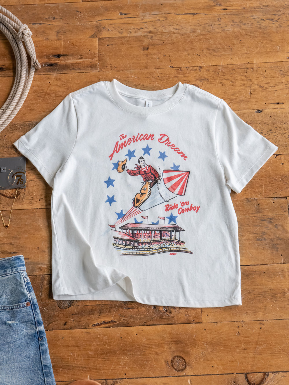 The American Dream tee by Savannah Sevens western life{&}style lies on a wooden floor, featuring a cowboy riding a guitar, blue stars, and “The American Dream” and “Ride em Cowboy” text. 100% cotton for comfort. Blue jeans and necklace nearby.