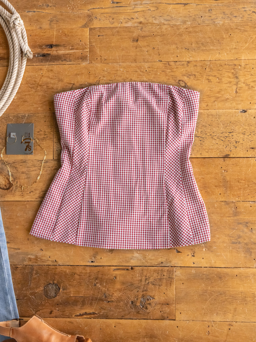 The Sanibel by Savannah Sevens western life{&}style is a red and white gingham strapless top in young contemporary sizing, crafted from a cotton blend and shown flat on wood with a metal branding iron and part of a leather boot nearby.