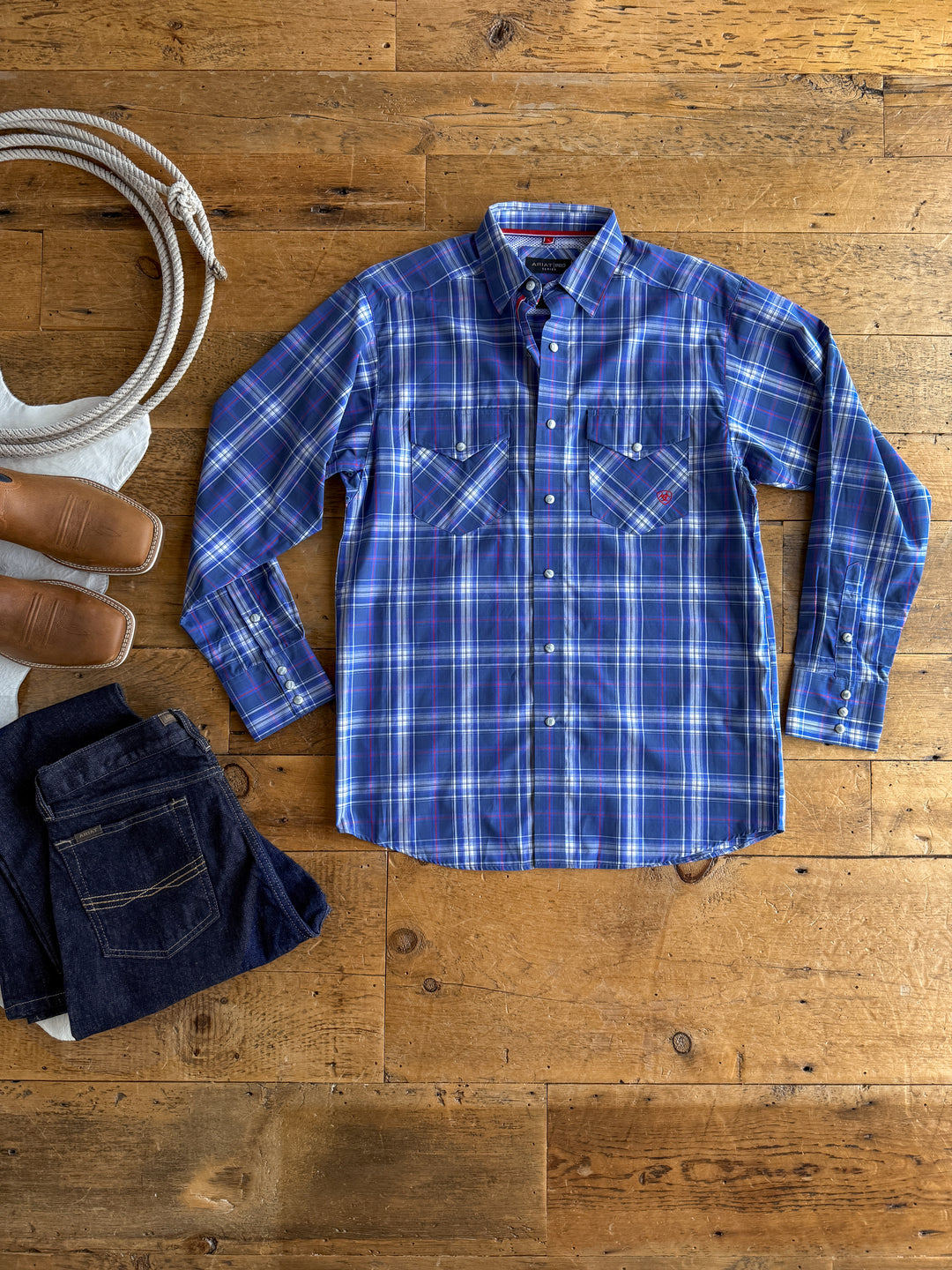 The Rockford men's blue long sleeve plaid shirt by Savannah Sevens western life&style is laid flat on a wooden floor, accompanied by jeans and brown boots.