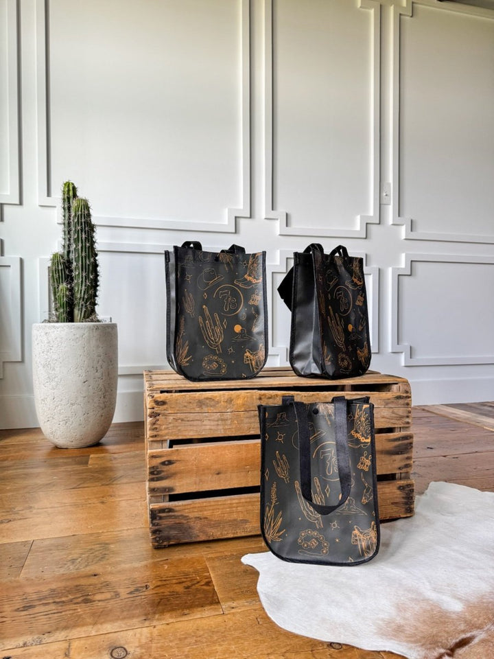 Three Savannah Sevens 7s {Life&Style} Reusable Bags with gold western toile designs are displayed on and beside a rustic crate in a bright room. Each bag features durable handles, perfect for stylish, sustainable use.