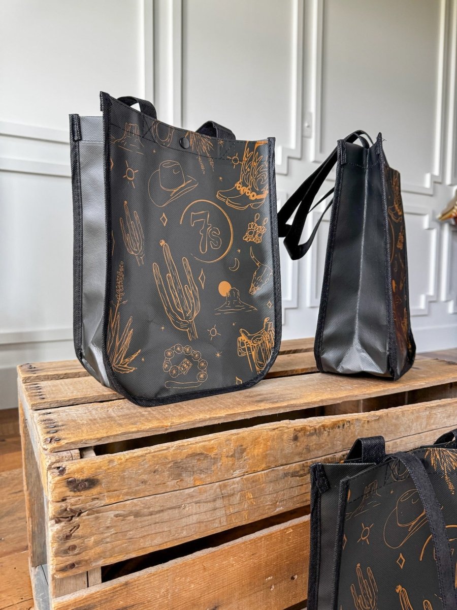 Three 7s {Life&Style} Reusable Bags by Savannah Sevens western life{&}style, featuring orange Southwestern designs—cacti, cowboy hats, and boots—are displayed on a wooden crate. Each black bag has durable handles for everyday use in a bright room.
