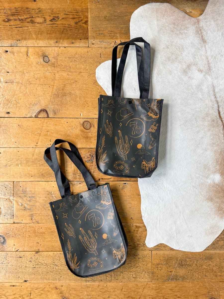Two black 7s {Life&Style} Reusable Bags by Savannah Sevens western life{&}style, with brown desert-themed doodles, sturdy handles, and western toile charm, rest on a wooden floor beside a white cowhide rug.