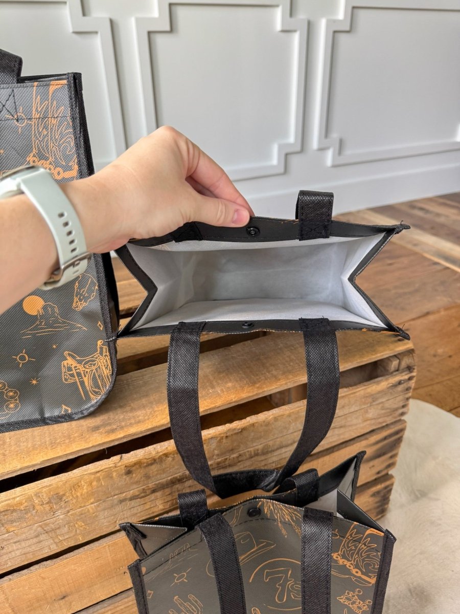 A hand holds open a Savannah Sevens western life{&}style 7s {Life&Style} Reusable Bag, featuring a black design with tan western toile and sturdy handles. Another identical bag and part of a wooden crate are seen on the wooden floor in the background.