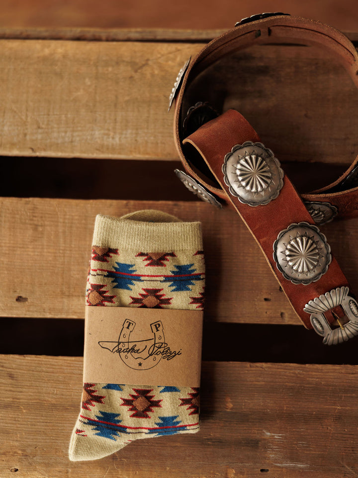 A pair of Savannah Sevens western life{&}style Rio Crew Socks with a vibrant Southwestern design and a brown belt with large silver conchos rest on a wooden surface.