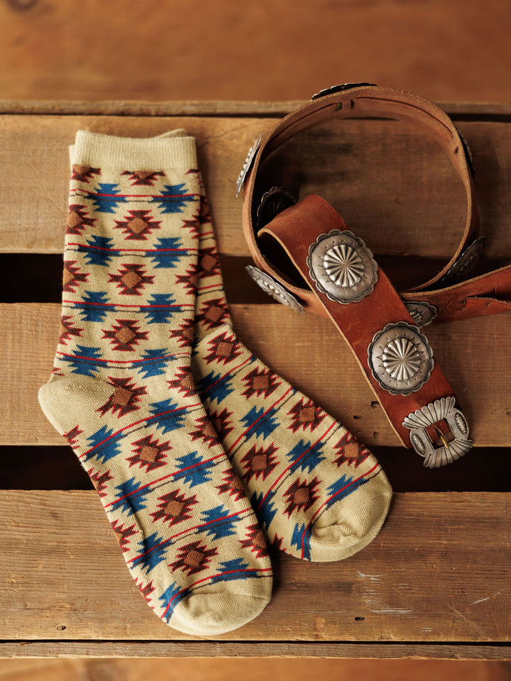 A pair of Savannah Sevens western life{&}style Rio Crew Socks in beige, red, and blue lies next to a brown leather belt with large silver conchos, both displayed on a wooden surface.