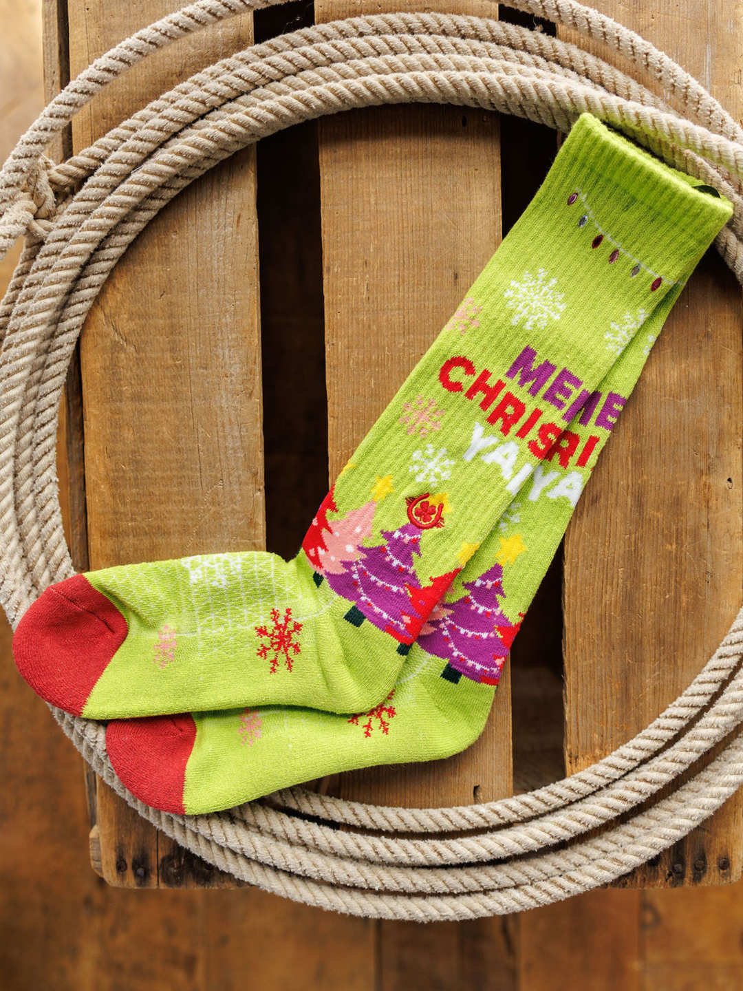 A pair of Merry Christmas Y’all Crew Socks (Green) by Savannah Sevens western life{&}style, featuring red toes, snowflakes, colorful Christmas trees, and "MERRY CHRISTMAS," displayed inside a coiled rope on wood.