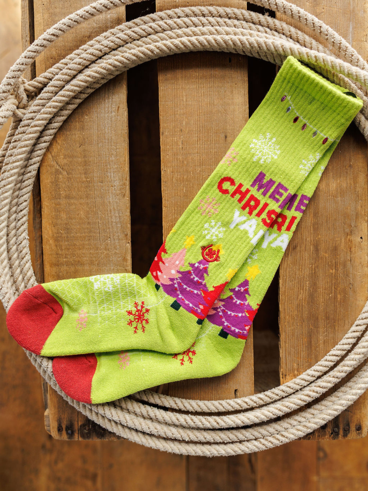 A pair of Merry Christmas Y’all Crew Socks (Green) by Savannah Sevens western life{&}style, featuring red toes, snowflakes, colorful Christmas trees, and "MERRY CHRISTMAS," displayed inside a coiled rope on wood.