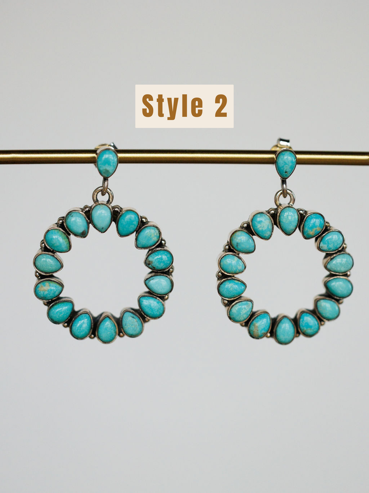 A pair of circular sterling silver Ocala Earrings by Savannah Sevens western life{&}style dangle from a gold rod, featuring Kingman turquoise teardrop stones. Above them, a label reads Style 2 in bold brown text.