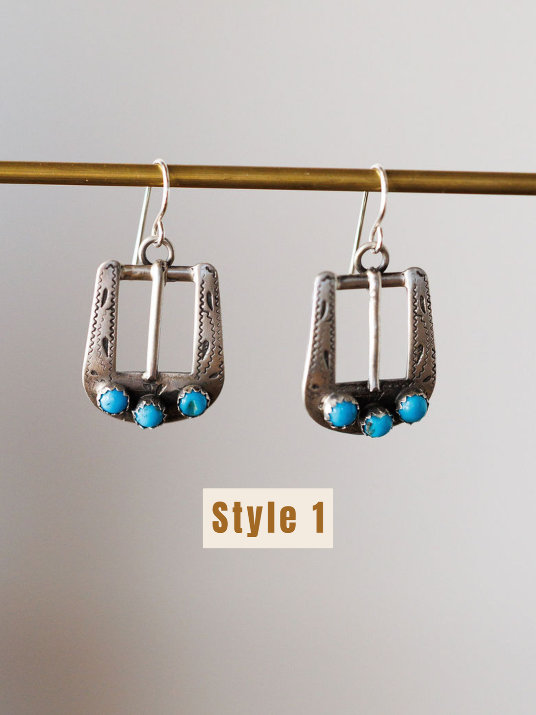 The Los Alamos Earrings by Savannah Sevens western life{&}style feature a rectangular sterling silver frame with ornate details and three small turquoise stones at the bottom. "Style 1" is shown below these turquoise earrings hanging on a rod.