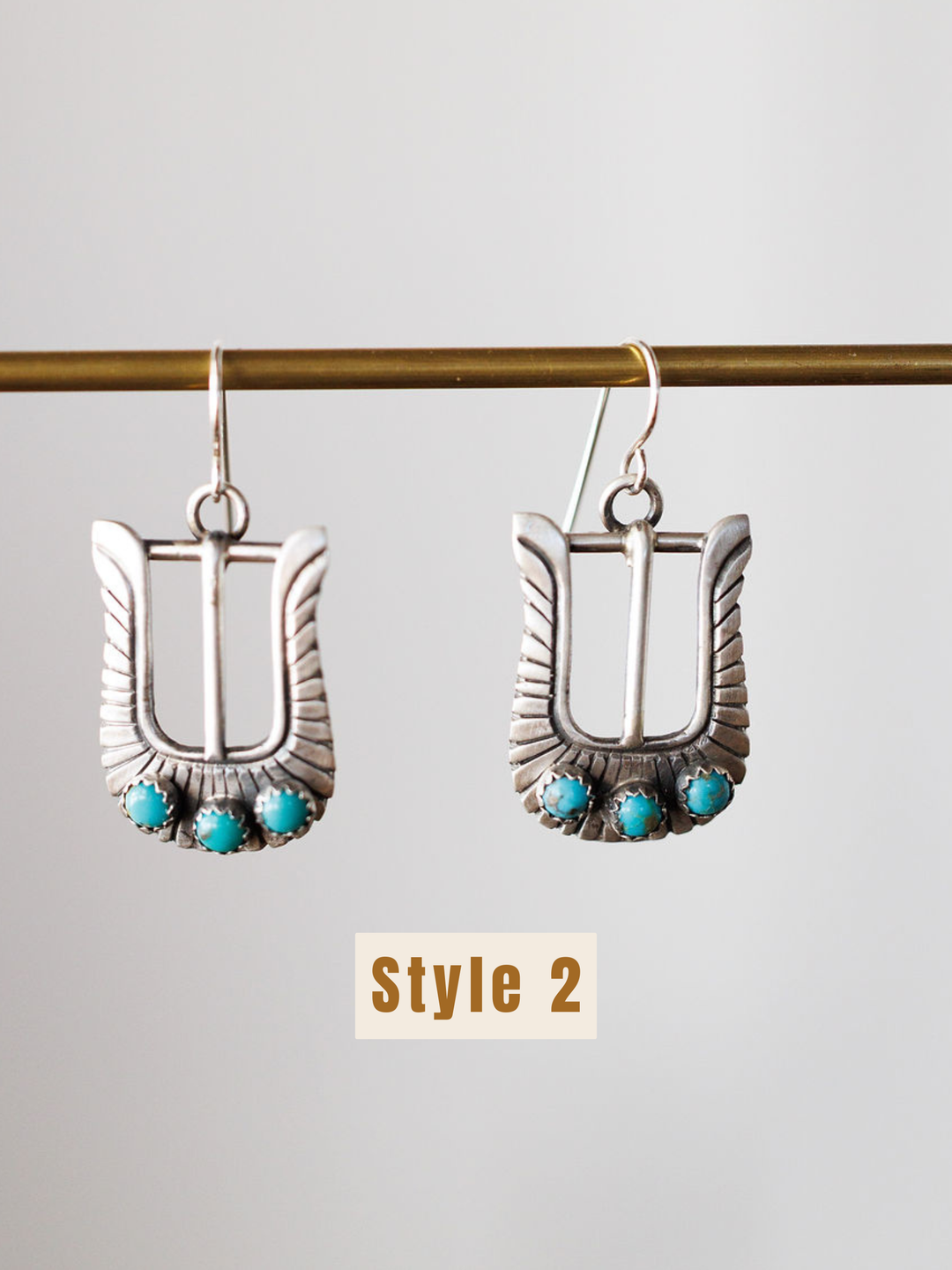 The Los Alamos Earrings by Savannah Sevens western life{&}style are sterling silver buckle-shaped earrings with three turquoise stones at the base, suspended from a horizontal rod. “Style 2” is shown below these unique turquoise earrings.