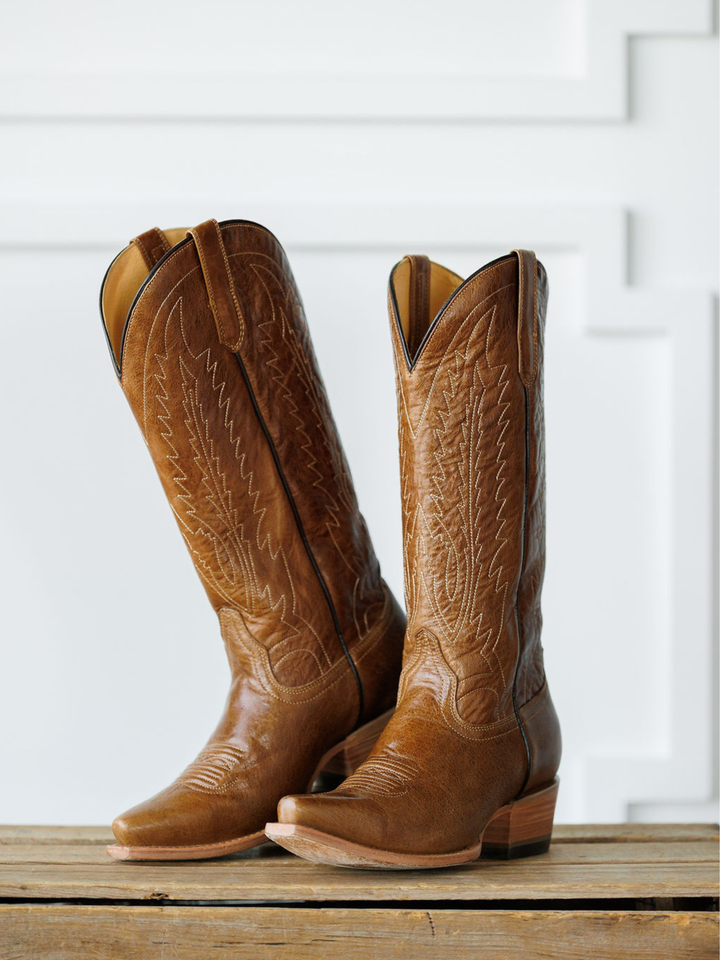 The Balcones boots from Savannah Sevens western life{&}style, tall and brown with intricate stitching, stand side by side on a wooden surface against a white decorative background. Made by Black Star. 