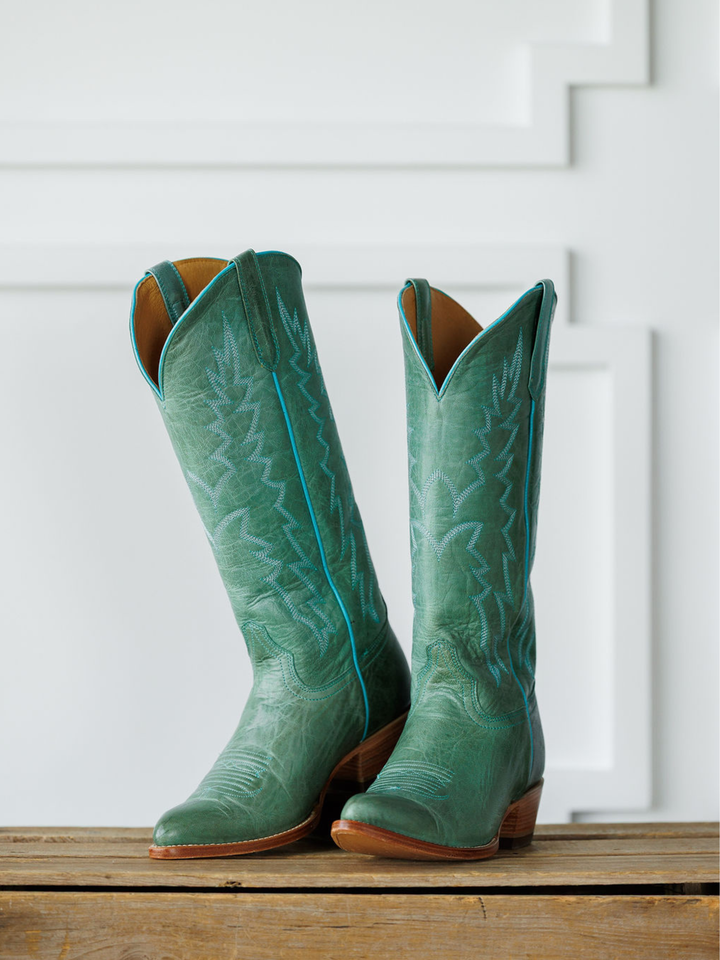 The Carico western boots by Savannah Sevens western life{&}style, featuring tall green turquoise leather with subtle embroidery and an almond toe, stand side by side on a wooden surface against a white paneled background. Made by Black Star. 