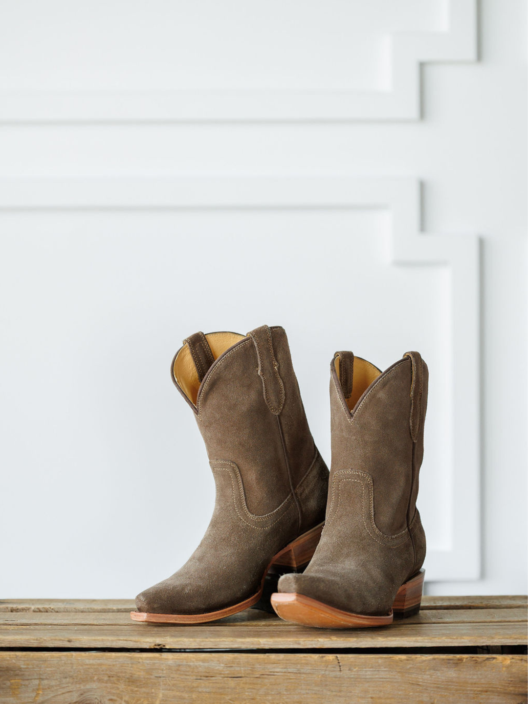 The Cordova booties by Savannah Sevens western life{&}style, crafted in brown rough out leather with wooden heels, rest on a rustic wooden surface before a white wall with geometric molding. Made by Black Star. 