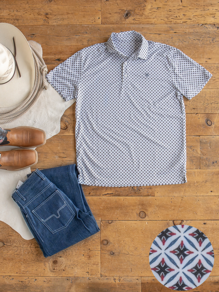 The Lyman men's short-sleeve polo by Savannah Sevens western life{&}style, featuring moisture wicking and a geometric red, blue, and black design, laid on wood floor beside blue jeans, tan boots, and a cowboy hat.