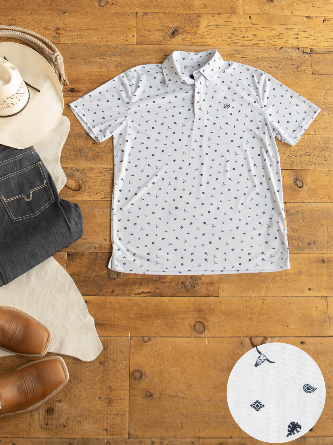 A Prescott Ariat Men's Western Graphic Polo by Savannah Sevens western life{&}style, featuring white fabric with black Western icons, is displayed flat on wood with a cowboy hat, jeans, boots, and a close-up of its moisture-wicking pattern.