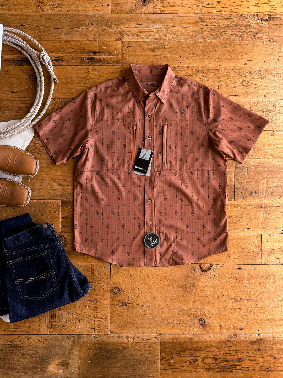 The Laramie men's short sleeve shirt by Savannah Sevens western life{&}style, shown in rust with a geometric pattern and tags attached, is laid flat beside brown sandals, a coiled rope, and folded dark blue jeans on a wooden floor.