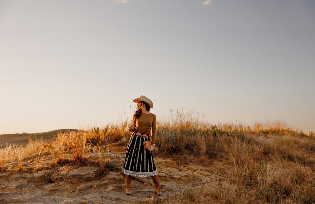 A woman in a cowboy hat and striped skirt stands on rocky ground surrounded by tall grass at sunset, gazing thoughtfully to her left.