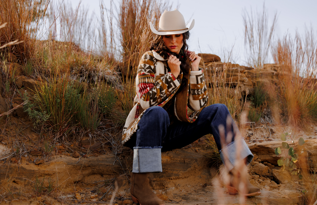 A person wearing a cowboy hat, patterned jacket, jeans, and boots sits on rocky ground among tall dry grass, looking down and holding their jacket collar. The scene captures a rugged western vibe.