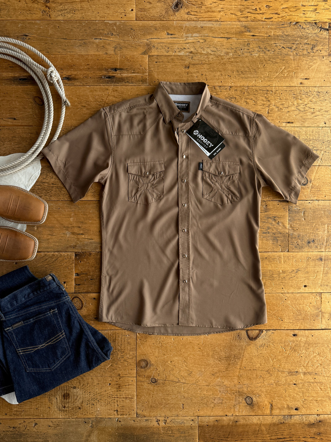 The Blaine men’s shirt by Savannah Sevens western life&style, in brown with short sleeves and tags, is laid flat on a wooden floor with blue jeans, tan slip-on shoes, and coiled rope. Features moisture-wicking for comfort.