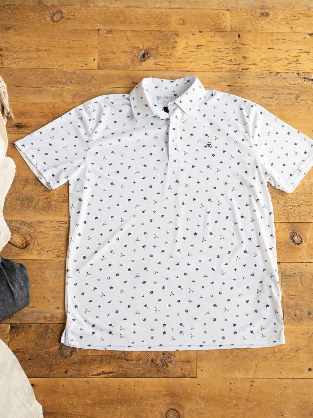 A Prescott Ariat Men's Western Graphic Polo by Savannah Sevens western life{&}style is laid flat on a wooden floor. The white short-sleeve shirt features sun protection, a collar, neckline buttons, and a small repeating navy blue pattern.