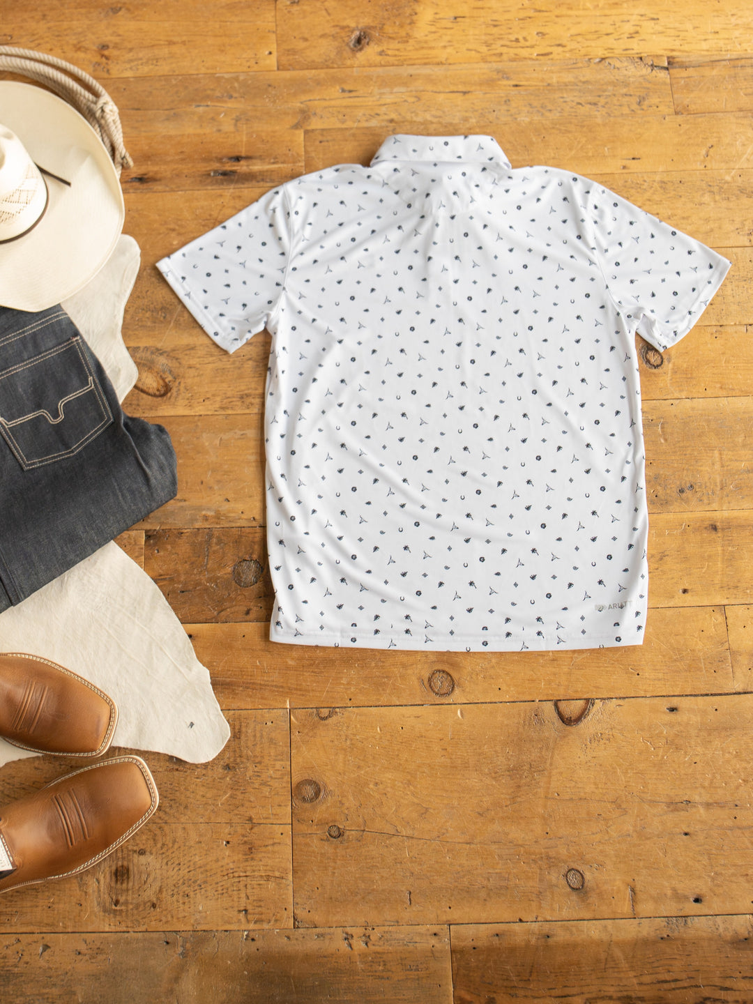 The Prescott Ariat Men's Western Graphic Polo by Savannah Sevens western life{&}style, a white short-sleeve moisture-wicking shirt with a black print, is styled flat with tan shoes, dark jeans, a beige hat, and light cloth—perfect for active days.