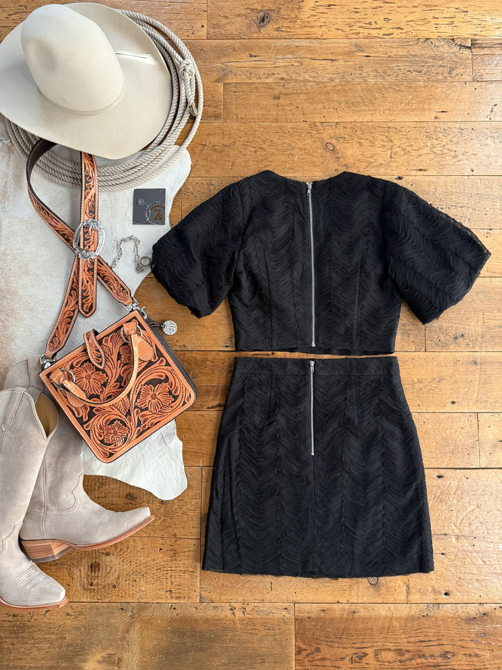 Black dress on a wooden surface with cowboy hat, boots, and leather bag.