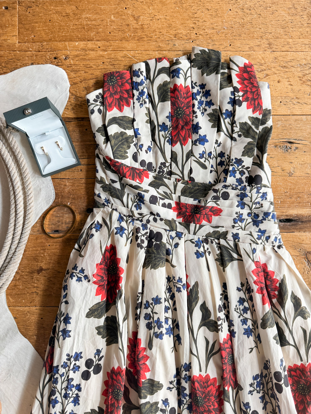 The Coppell Cream Floral Strapless Dress by Savannah Sevens western life{&}style is displayed on a wooden surface alongside a boxed pair of earrings, a silver bracelet, a white hat, and a beige rope.