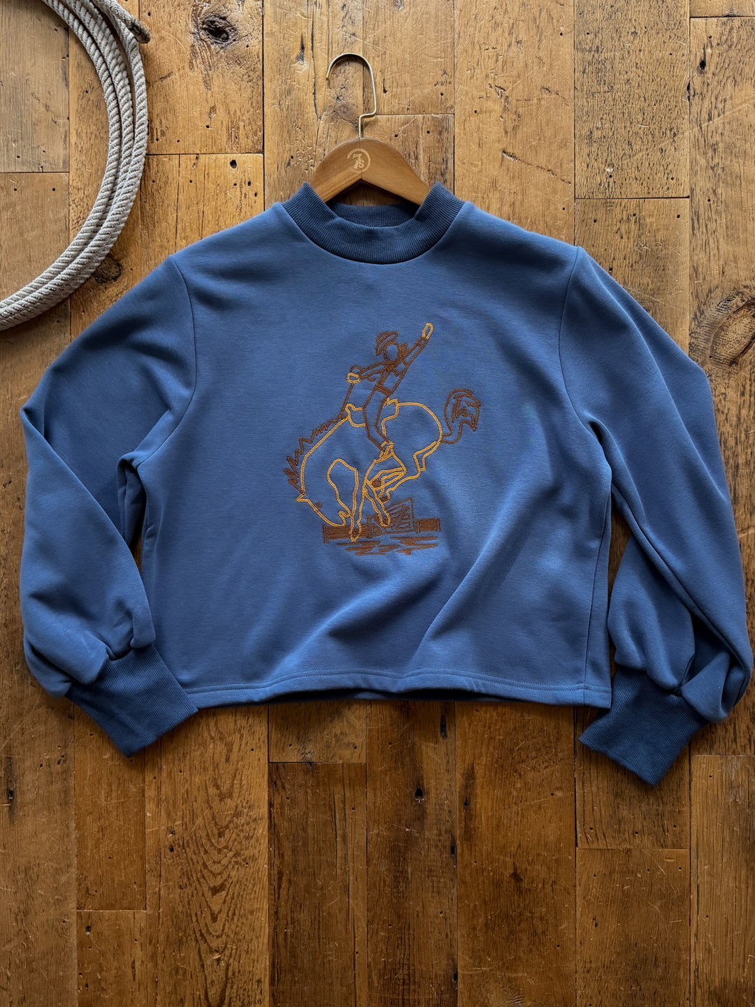 Savannah Sevens western life&style Saddle Bronc Slate Blue Embroidered Pullover, a slate blue fleece-lined mock-neck featuring a gold chain-stitch embroidered graphic of a cowboy riding a bucking bronc.