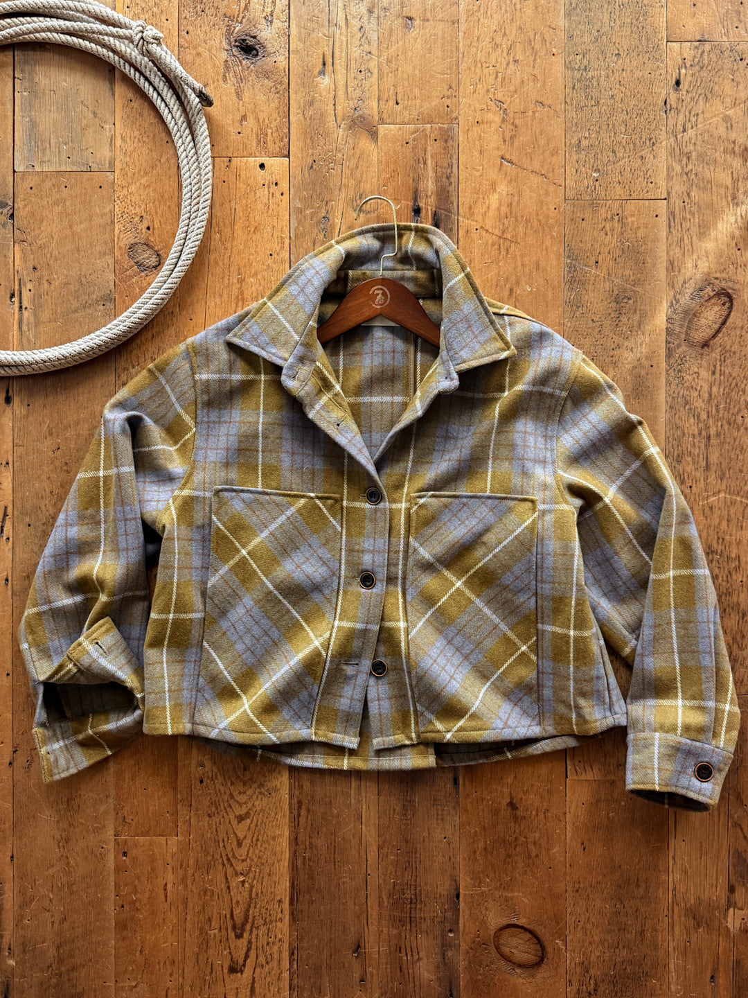 Relaxed-fit olive green and grey plaid Taloga Plaid Wool Blend Jacket by Savannah Sevens western life&style, featuring large patch pockets and hanging on a wooden hanger against a wood plank background.