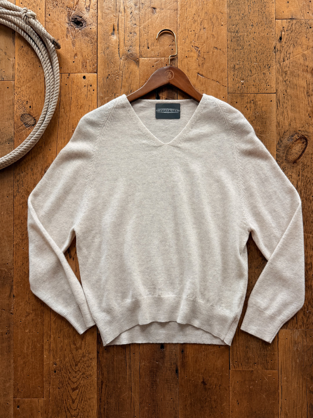 Avant Oatmeal Wool Cashmere Sweater in oatmeal-colored wool cashmere hanging on a wooden hanger against a wood floor background.