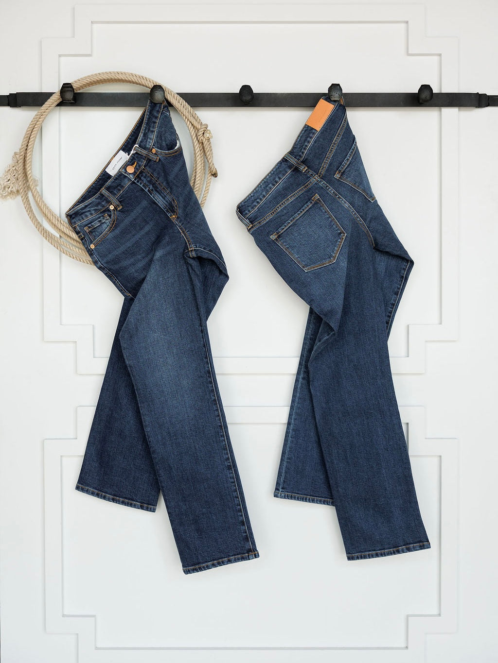 Two pairs of The Camryn Premier Rise Slim Straight Leg Jeans by Savannah Sevens western life{&}style hang side by side on a black wall-mounted rod with a coiled rope, set against a white decorative wall.