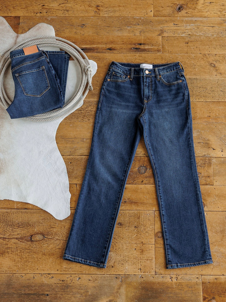 The Camryn Premier Rise Slim Straight Leg Jean by Savannah Sevens western life{&}style is displayed flat on a wooden floor beside a folded pair of jeans and coiled rope atop a white cowhide rug.
